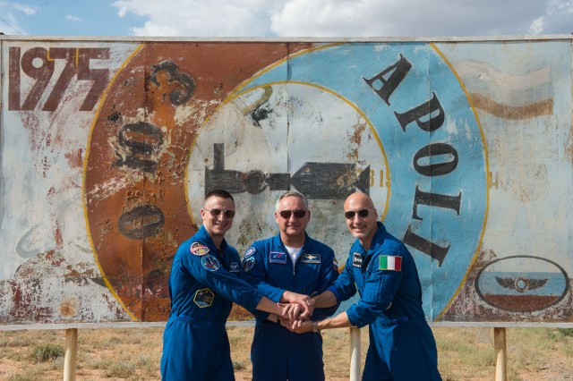 jsc2019e038379 (July 5, 2019) --- At the Baikonur Cosmodrome in Kazakhstan, Expedition 60 crewmembers Drew Morgan of NASA (left), Alexander Skvortsov of Roscosmos (center) and Luca Parmitano of the European Space Agency (right) pose for pictures July 5 in front of a mural bearing the insignia of the 1975 Apollo-Soyuz mission. They will launch July 20 on the 50th anniversary of humanity’s first landing on the moon during Apollo 11 on the Soyuz MS-13 spacecraft from the Baikonur Cosmodrome for a mission on the International Space Station. Credit: Andrey Shelepin/GCTC