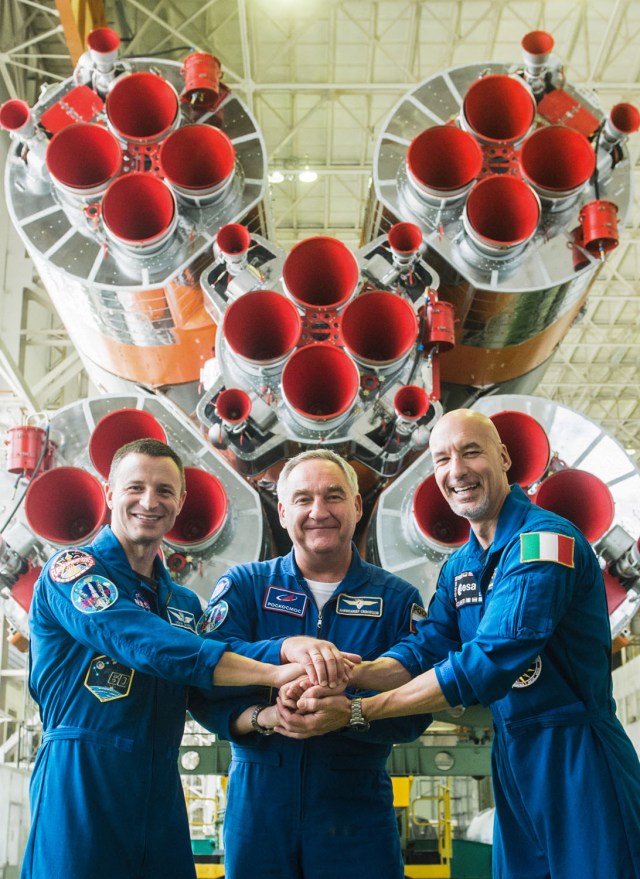 jsc2019e039425 (July 16, 2019) --- In the Integration Building at the Baikonur Cosmodrome in Kazakhstan, Expedition 60 crewmembers Drew Morgan of NASA (left), Alexander Skvortsov of Roscosmos (center) and Luca Parmitano of the European Space Agency (right) pose for pictures in front of the first stage engines of their Soyuz booster July 16 as part of pre-launch preparations. They will launch July 20 on the Soyuz MS-13 spacecraft from the Baikonur Cosmodrome for a mission on the International Space Station. Credit: Andrey Shelepin/GCTC
