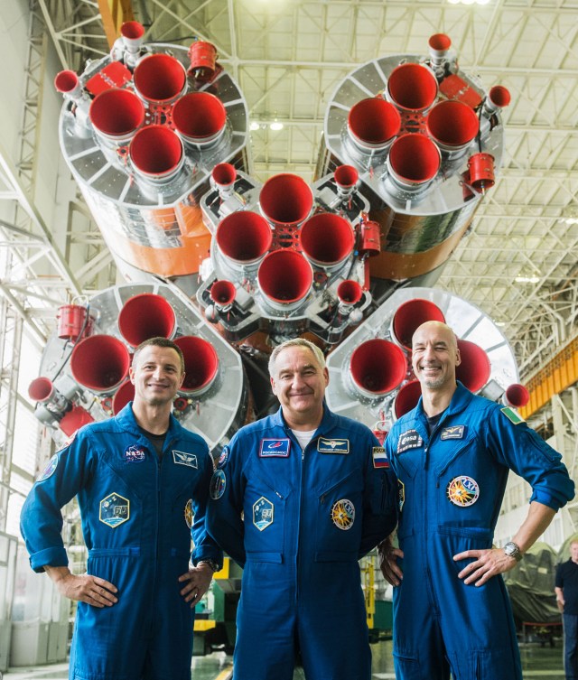 jsc2019e039429 (July 16, 2019) --- In the Integration Building at the Baikonur Cosmodrome in Kazakhstan, Expedition 60 crewmembers Drew Morgan of NASA (left), Alexander Skvortsov of Roscosmos (center) and Luca Parmitano of the European Space Agency (right) pose for pictures in front of the first stage engines of their Soyuz booster July 16 as part of pre-launch preparations. They will launch July 20 on the Soyuz MS-13 spacecraft from the Baikonur Cosmodrome for a mission on the International Space Station. Credit: Andrey Shelepin/GCTC