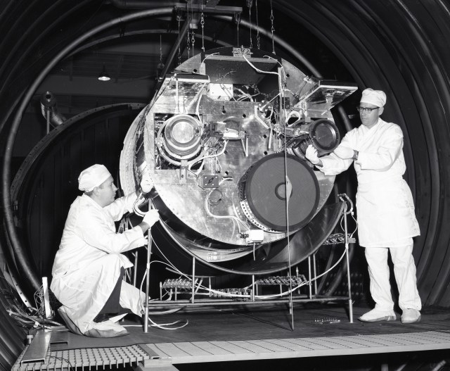 In this black-and-white photo, two male NASA employees wearing white protective gear work on a large piece of space hardware with thrusters attached. They are inside a large vacuum chamber.