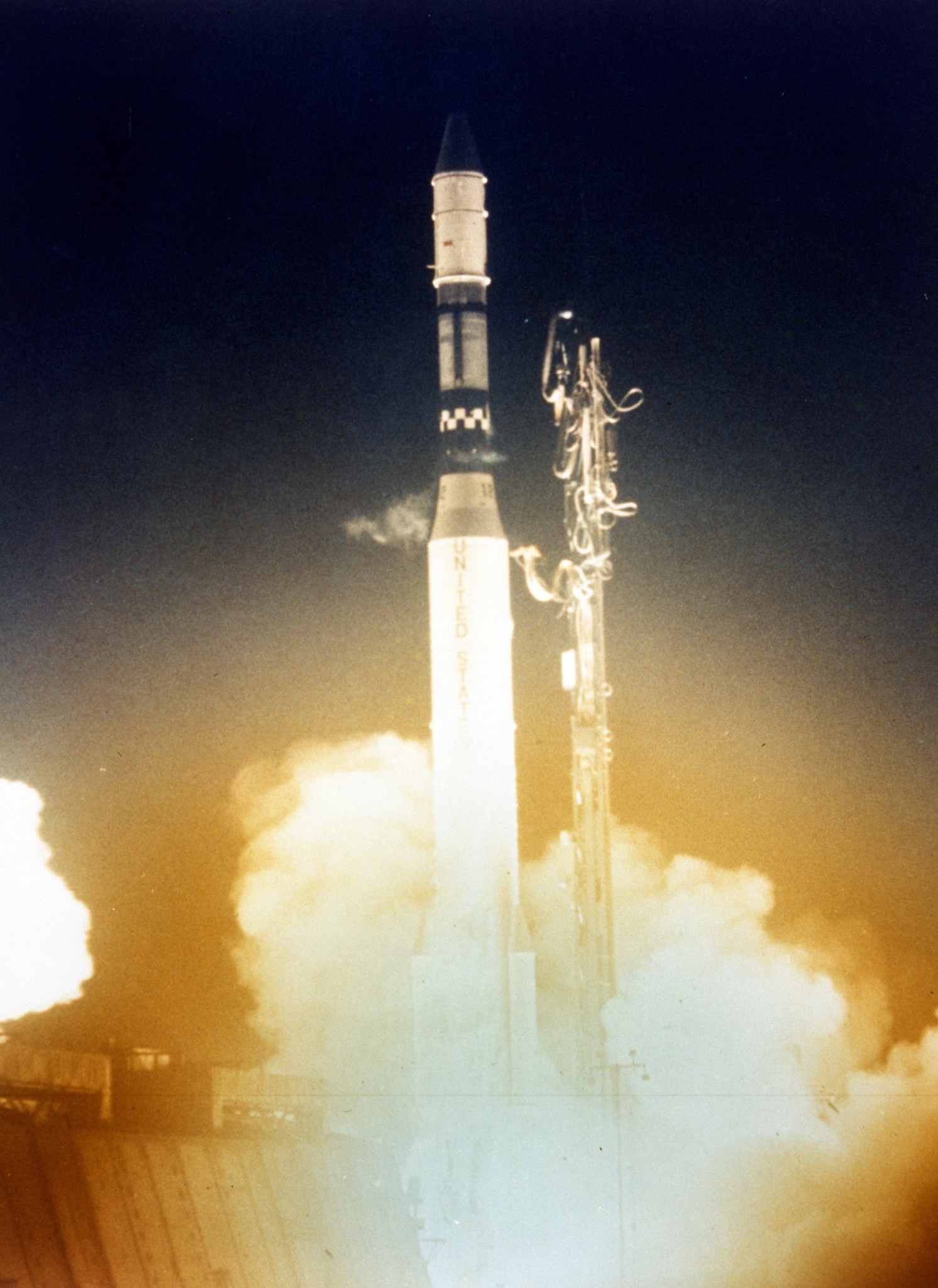 A rocket launches and smoke billows up from below. The background of the photo is dark blue and orange light can be seen coming from below the rocket.