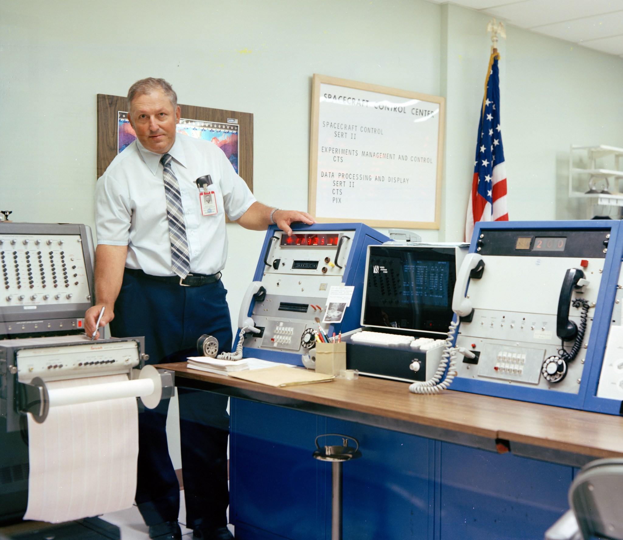 A man wearing a tie and a NASA employee badge stands in a control room next to various computers and consoles. He looks at the camera, and behind him is an American flag and a sign that says, “Spacecraft Control Center” and “SERT II.”