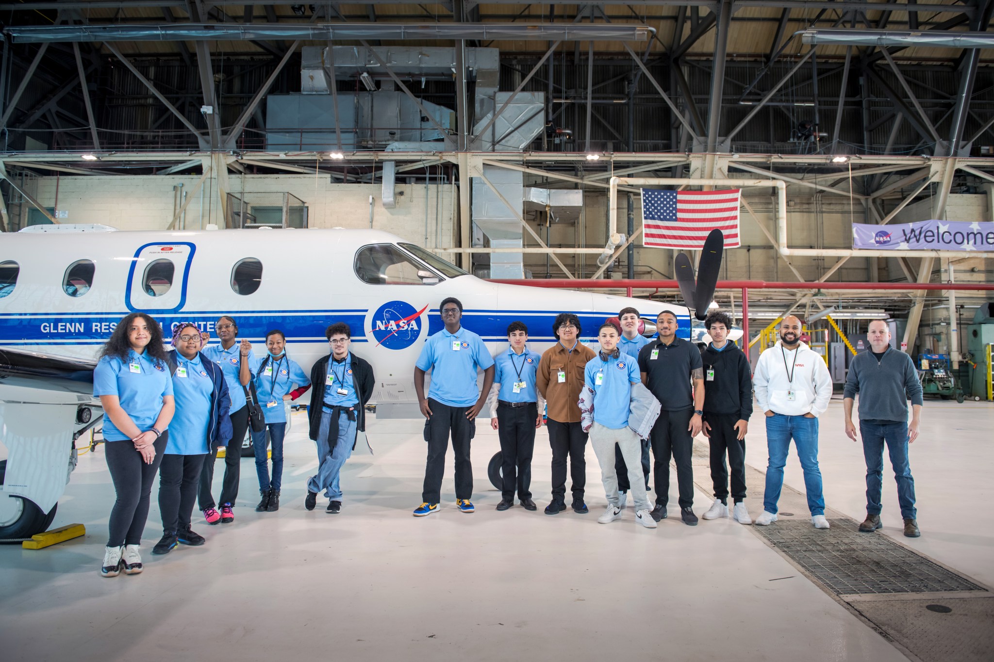 A group of high school students dressed in blue shirts and dark pants pose in front of a small airplane in a hangar at NASA’s Glenn Research Center.