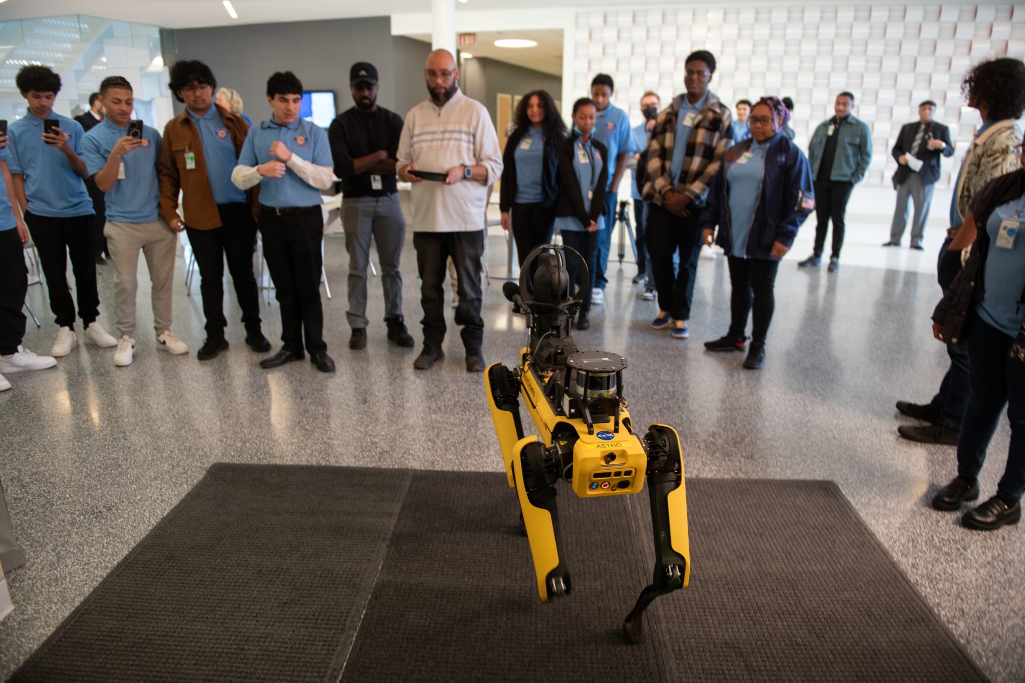 A group of high school students dressed in blue shirts and dark pants observe a demonstration with a yellow robotic dog in a large hallway . A man holds a remote control in his hands.