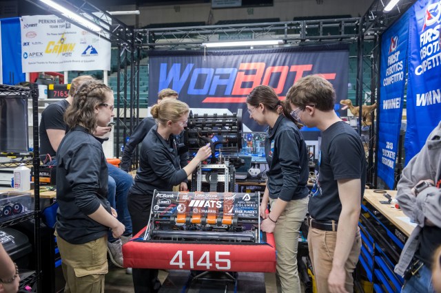 A group of six team members gather around their robot, looking at it and adjusting mechanisms. A banner with the name “WorBots” is in the background.