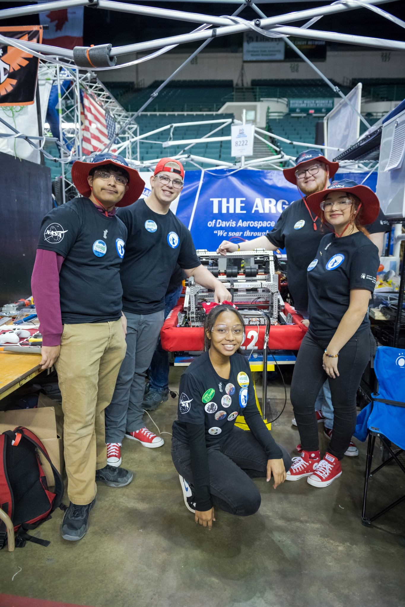 Four students stand and one student crouches down on the floor in front of their robot. The team’s name is partially shown on a banner behind them.