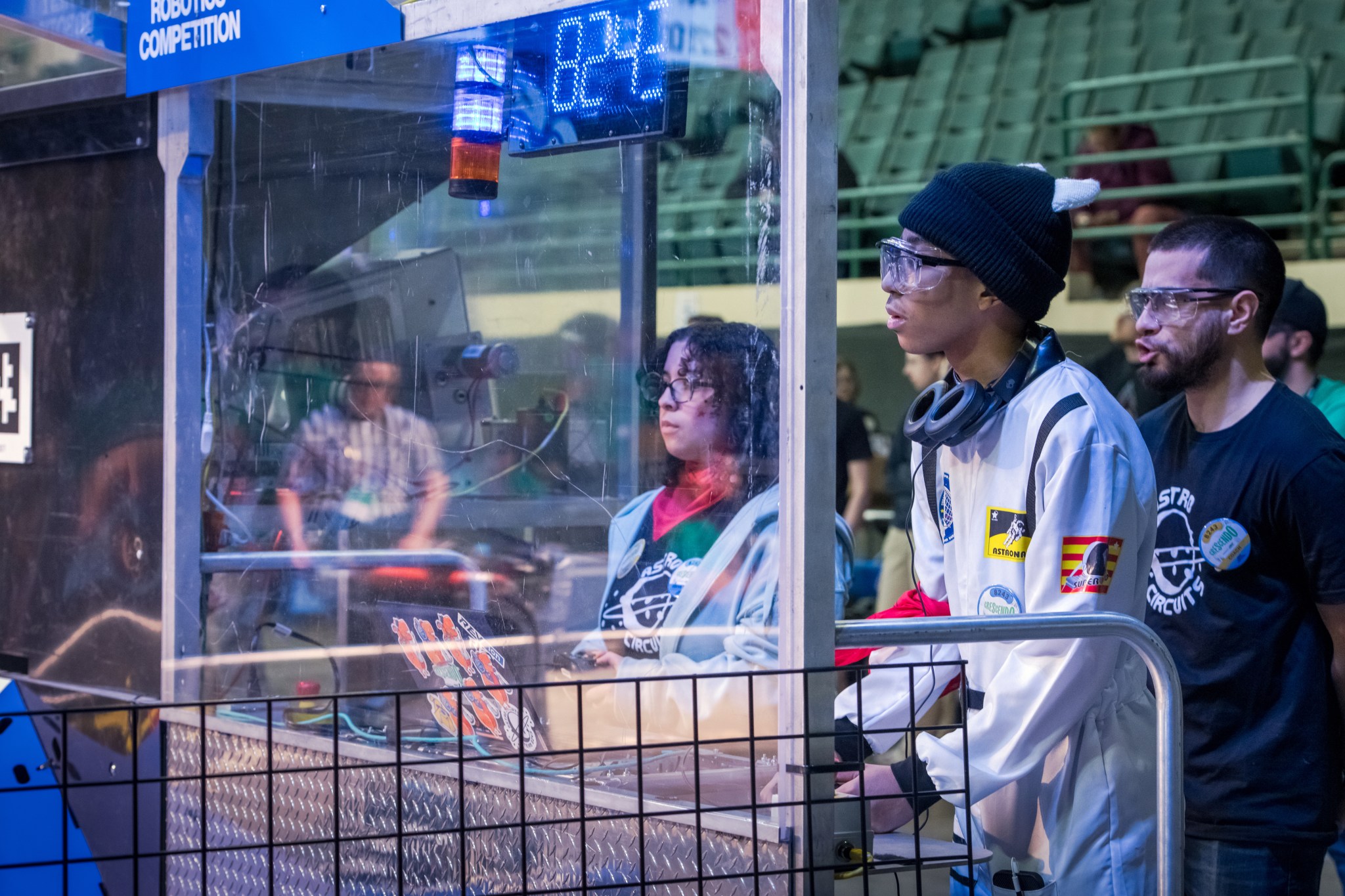Two team members stand behind a plexiglass screen and work controls for their robot. One person cheers them on from behind.