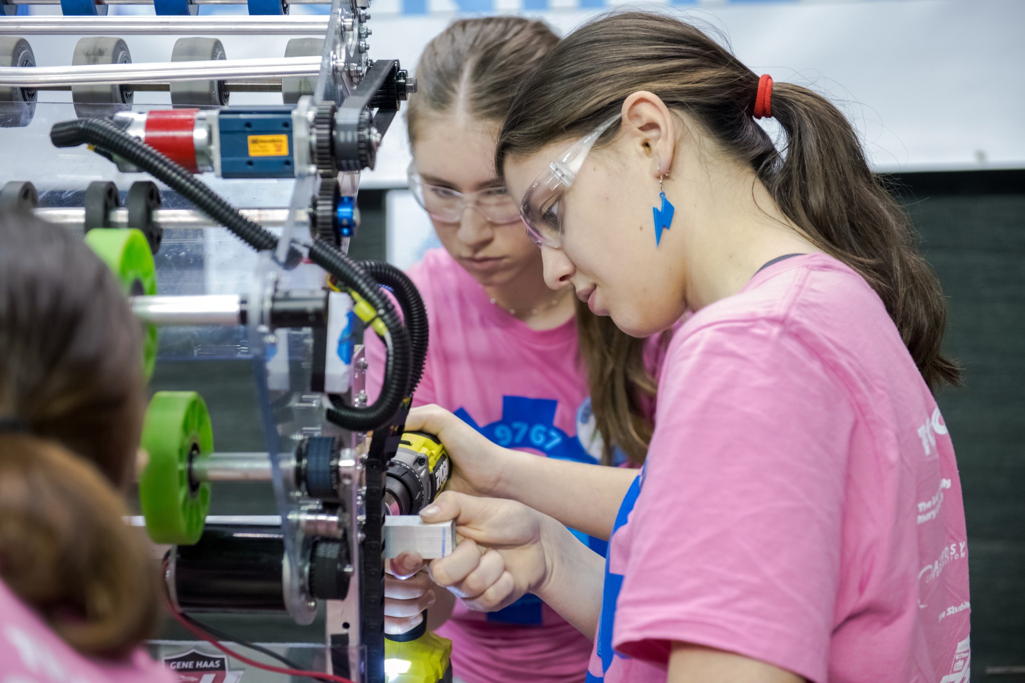 Two teenage girls in pink T-shirts adjust their robot.