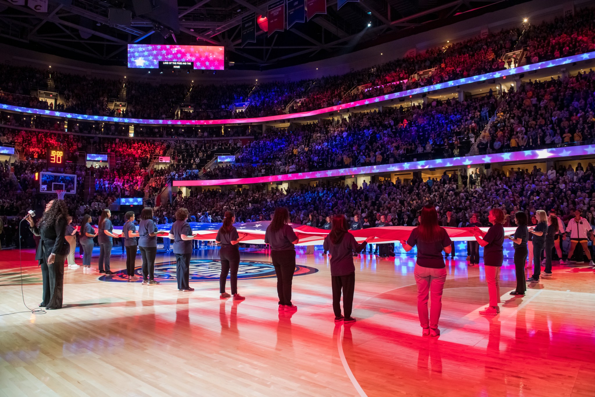 Several women hold ends of a large American flag on center court inside Rocket Mortgage FieldHouse. A woman stands at left singing the National Anthem. Red and blue lights illuminate the stage and stands full of guests. 