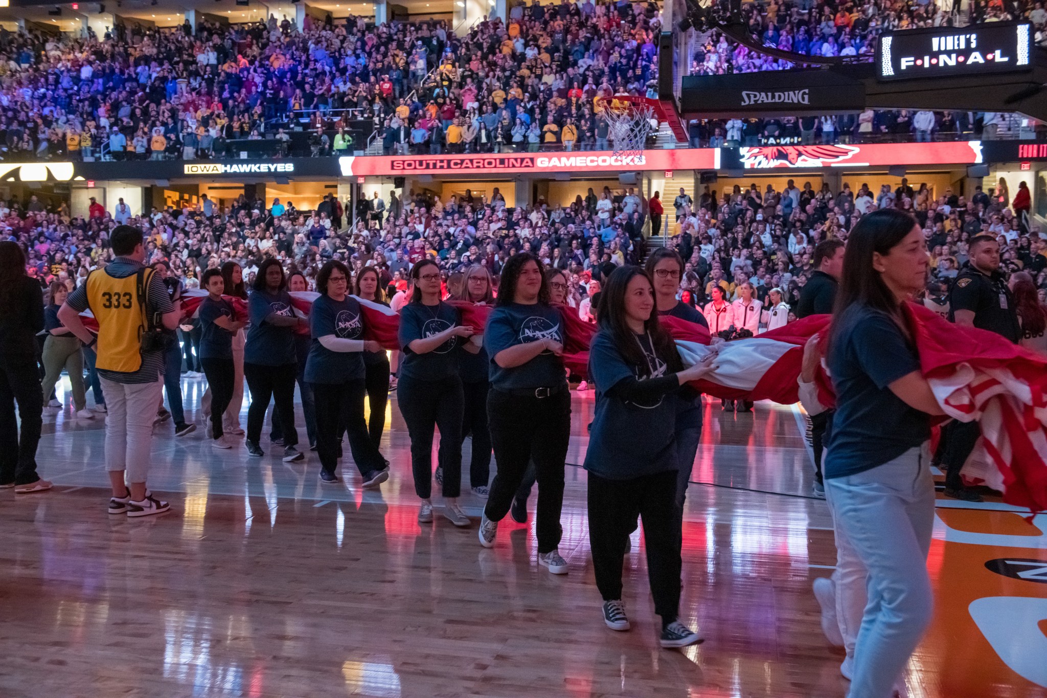 Several women line up in two rows while holding the large folded American flag. They walk off the court. The stands are full of guests. 