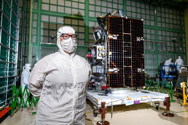 A woman stands on the left side of the image, seen from the waist up. She wears a white clean suit that covers her arms, torso, and over her head. She also has on a white mask that covers her nose and mouth and blue latex gloves. Behind her and to the right in the image is a large, rectangular piece of equipment - the PACE satellite. The side of the satellite visible is black and reflects light off it. Behind the woman and PACE is a large wall that has a green gridded pattern on it.