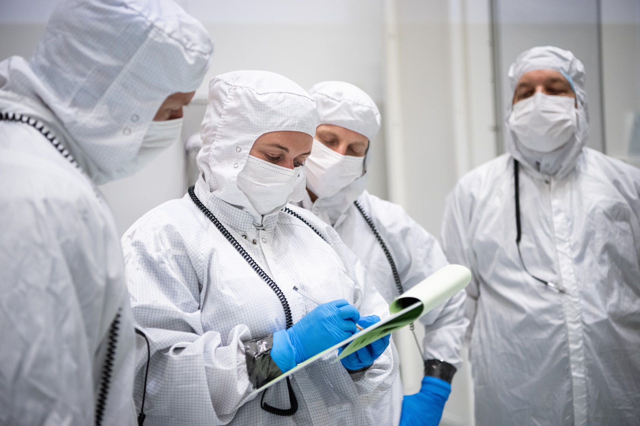 Four people stand in a white room wearing white clean suits with blue gloves. They have black cords draped over their shoulders and the woman in the middle is holding a note pad and pen, the other three people are looking at what she's writing. 