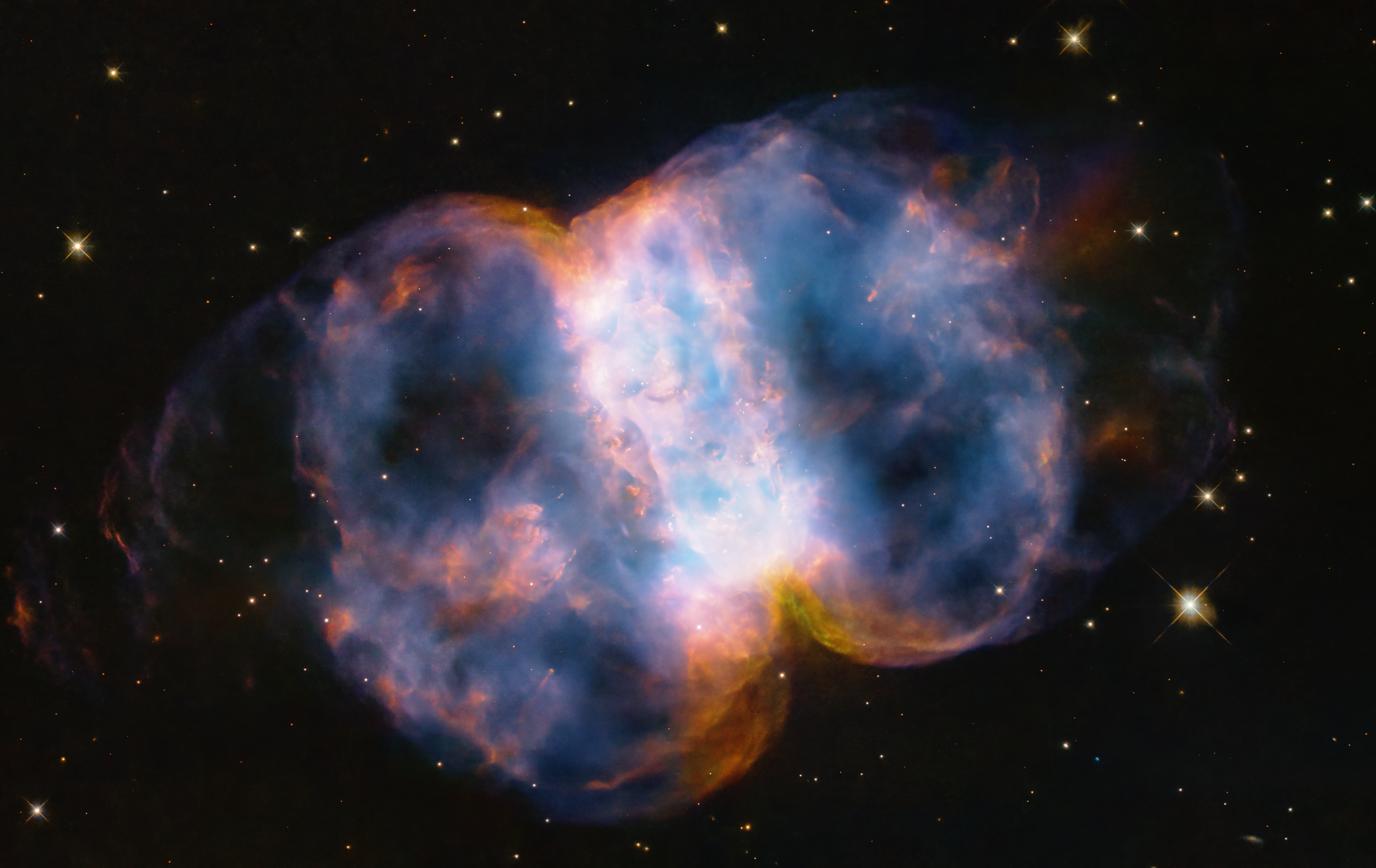 Taking up most of the image, is a multi-colored nebula in shades of blue, pink, yellow, orange, purple, and white. It appears as two translucent orbs attached by a white band.