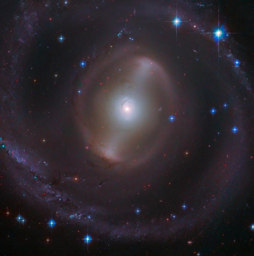 A bright, white galactic core shines near the center of the image with a faint bar of stars extending from it, diagonally to the right. Faint, hazy spiral arms encircle the core, with several distant stars and bright blue foreground stars with diffraction spikes scattered throughout the image, all against black space.
