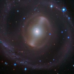 A bright, white galactic core shines near the center of the image with a faint bar of stars extending from it, diagonally to the right. Faint, hazy spiral arms encircle the core, with several distant stars and bright blue foreground stars with diffraction spikes scattered throughout the image, all against black space.