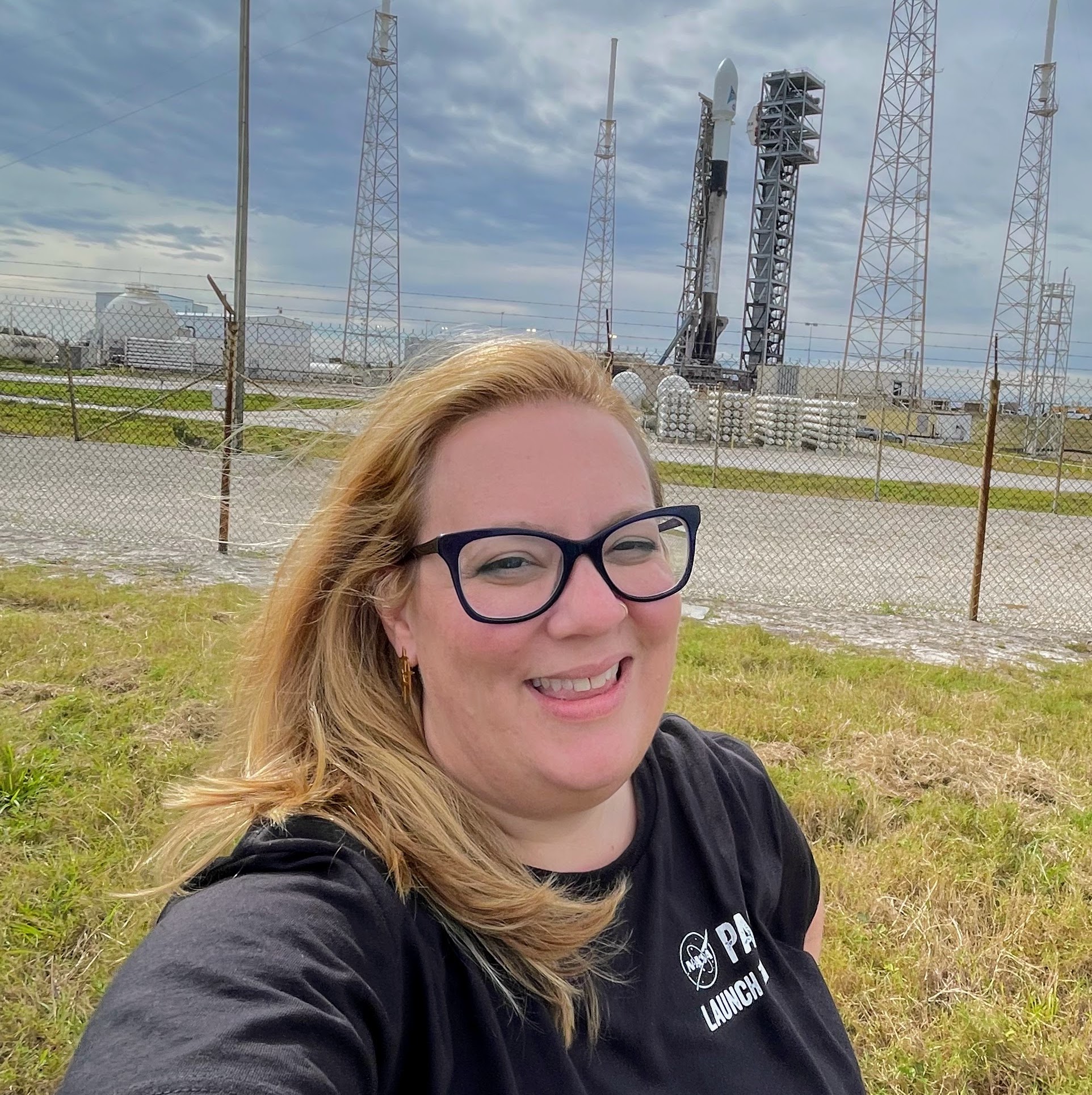 Selfie of Veronica Pinnick standing in front of a green lawn. She is smiling at the camera and wearing black glasses and a black shirt. Behind her, a Falcon 9 rocket is vertical on the launchpad.