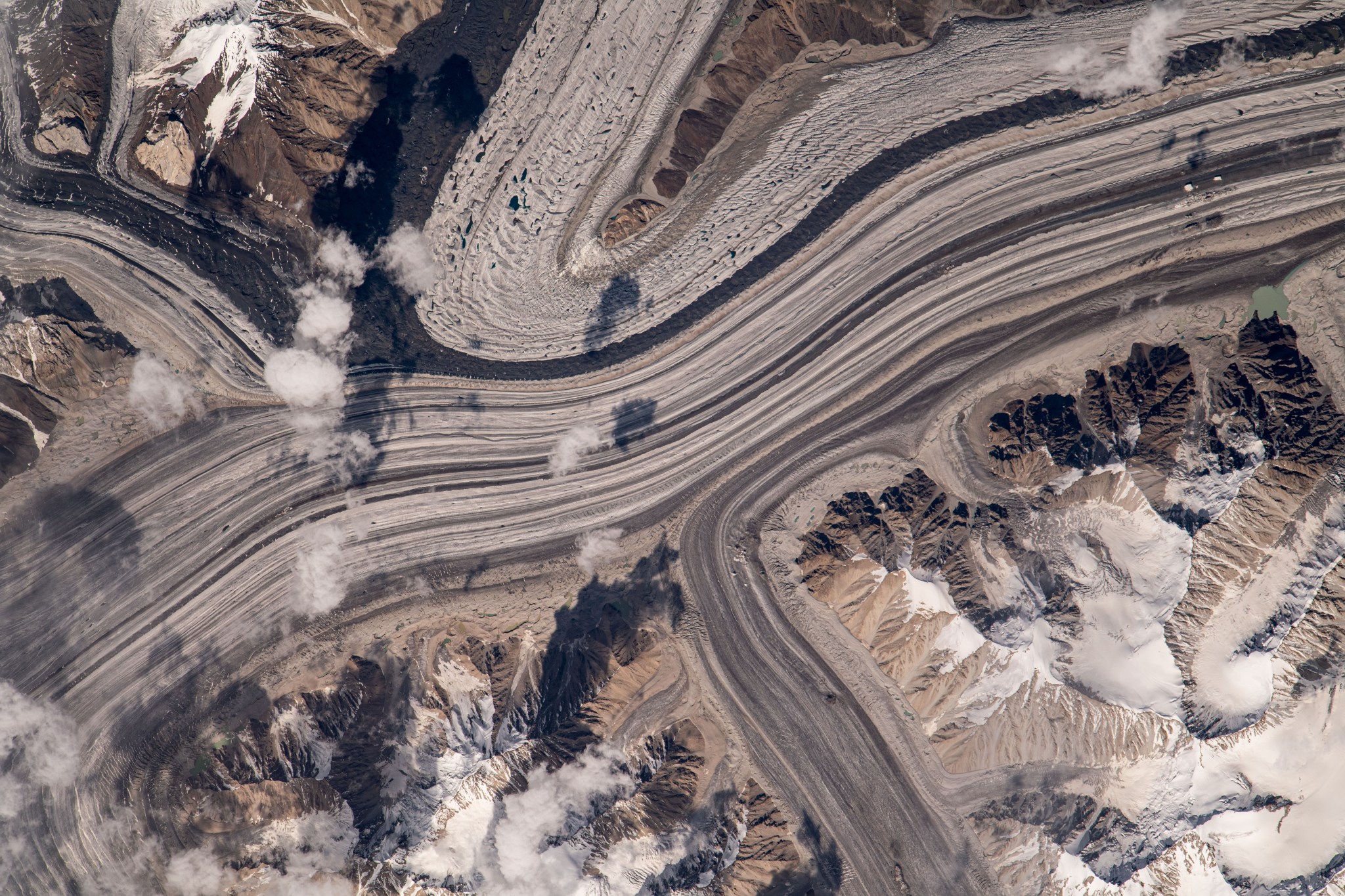 A view of mountains from the International Space Station. There are lines in various shades of gray that look like highways - these are paths carved by glaciers moving through the mountain range. Several peaks around the lines are untouched. A few clouds dot the sky at middle left and bottom left.