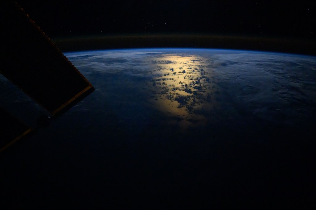 The Sun's glint beaming off the South Pacific Ocean near the coast of Chile just after an orbital sunrise and a slight airglow crowning Earth's horizon are photographed from the International Space Station. At top, are star fields including a cluster of stars in the constellation of Cancer and a portion of the Lynx constellation.