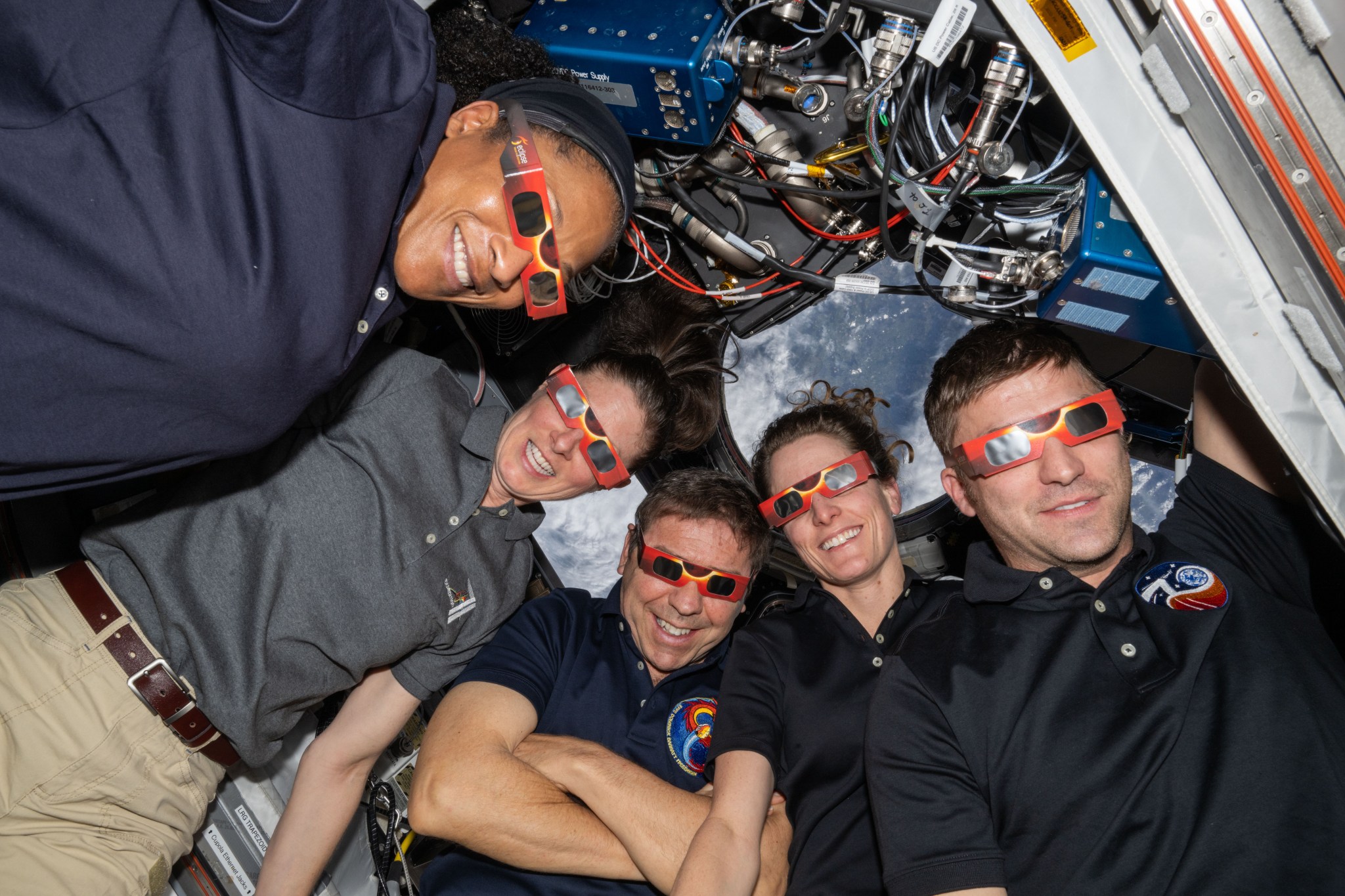 Five NASA astronauts, three women and two men, pose for a photo together while wearing eclipse glasses. The eclipse glasses are rectangular and have two dark lenses.