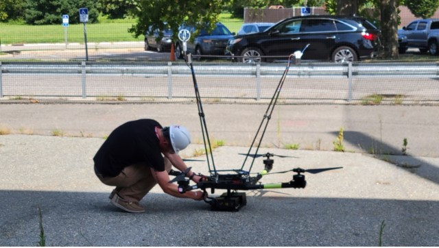 Jake Revesz, an electronic systems engineer at NASA Langley Research Center, is pictured here prepping a UAS for flight. Jake is kneeling on pavement working with the drone. He is wearing a t-shirt, khakis, and a hard hat.
