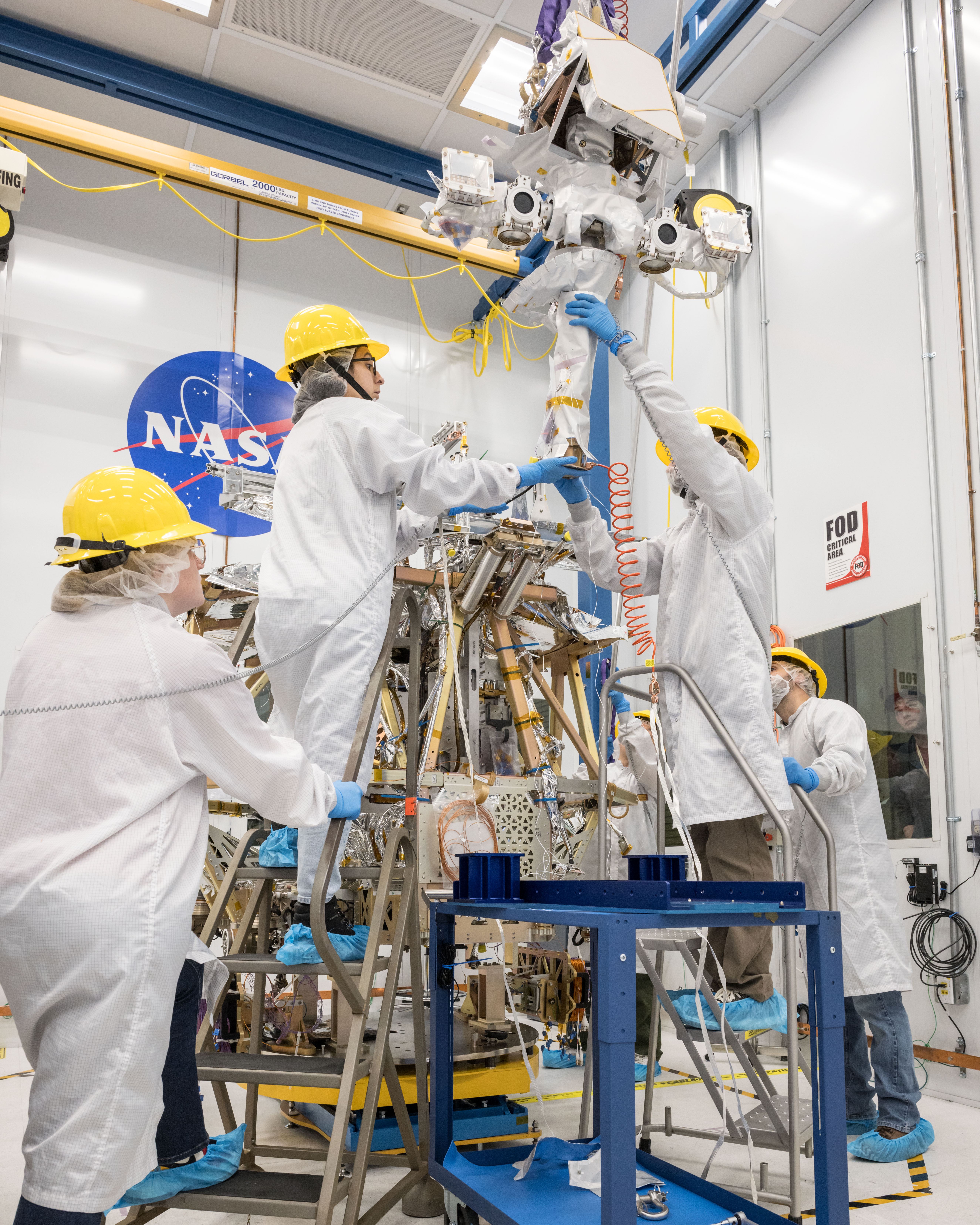 Four engineers in white lab coats, yellow hard hats, and blue gloves and shoe covers surround a metal rover. Two engineers stand on stepladders as they fit a mast with attached instruments onto the rover.
