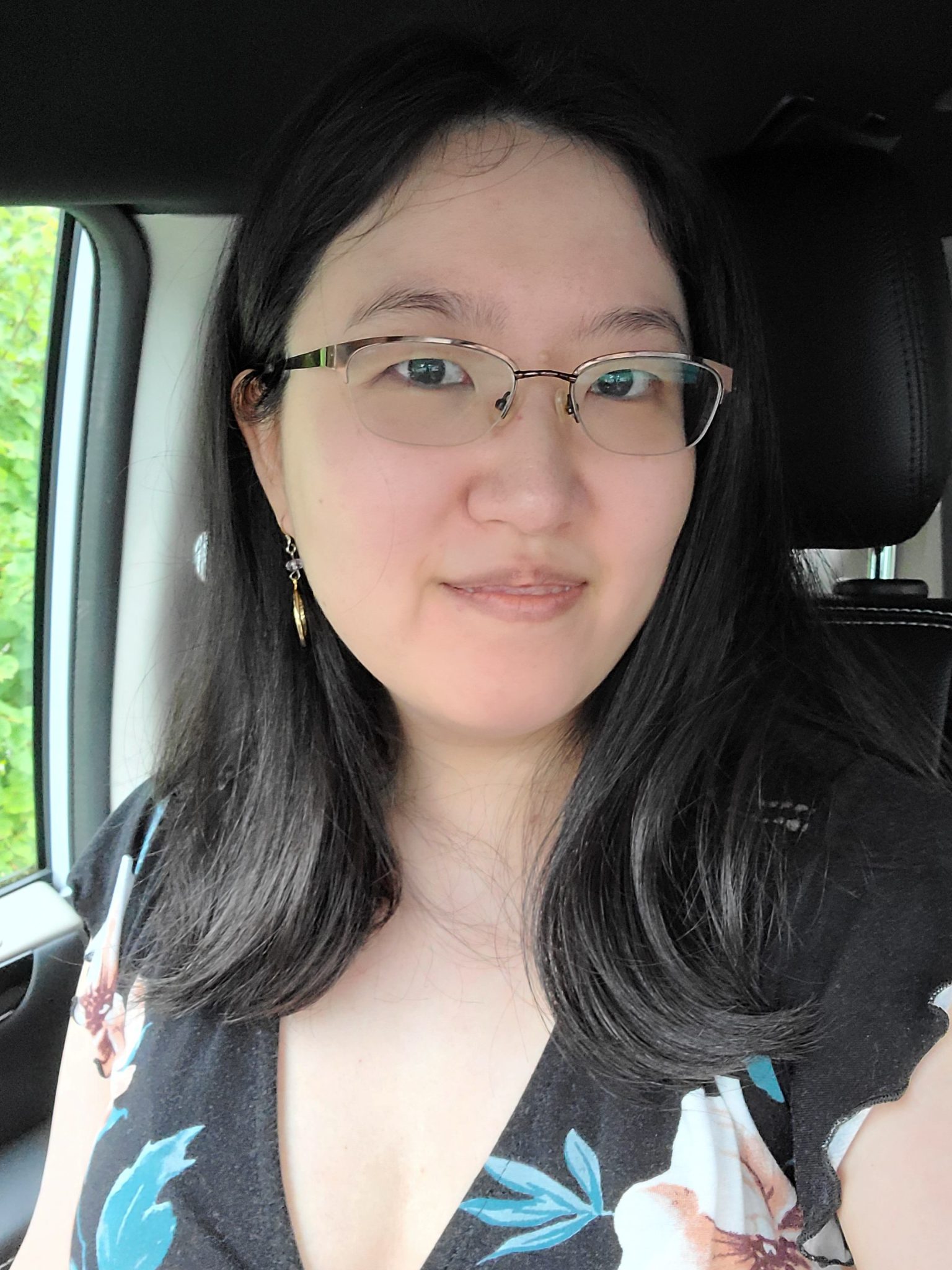  Kiyun Kim takes a selfie, smiling, in a car wearing a black, teal and peach floral top. They have shoulder length black hair and wear glasses.