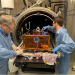 Photo of two men in blue lab jackets working on a copper colored box with wires