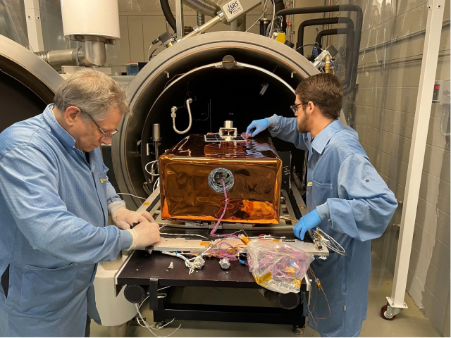 Photo of two men in blue lab jackets working on a copper colored box with wires