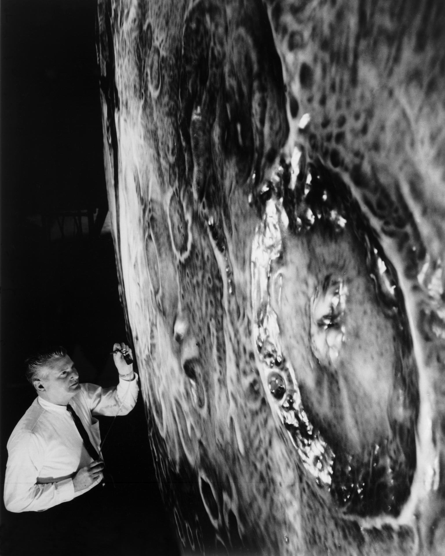 In this black-and-white photo, a man in a white shirt and dark tie uses an airbrush to paint on a large model that looks like the surface of the Moon. A crater is in the foreground, emphasizing the large size of the Moon model.