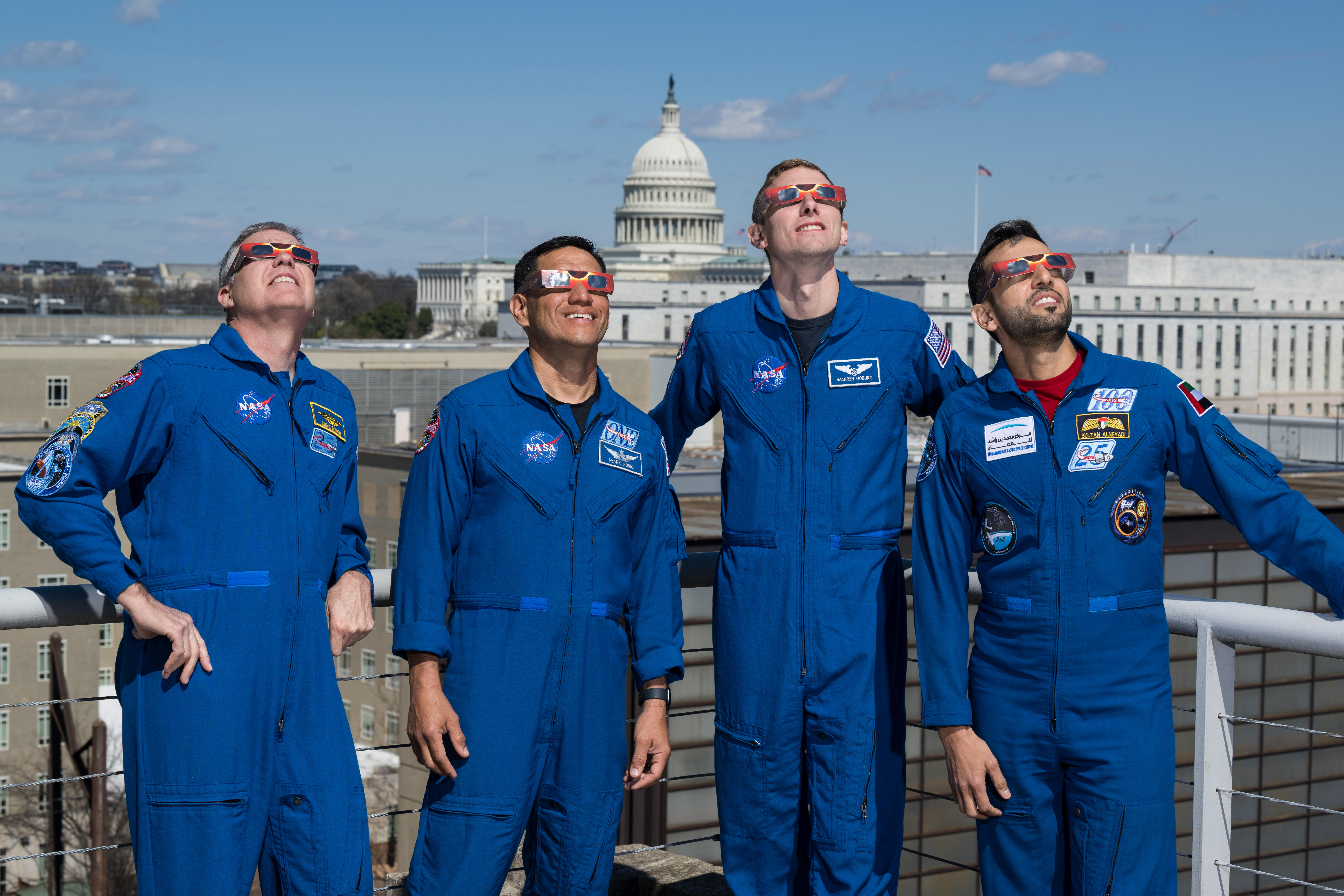 Four men wearing red, rectangular eclipse glasses and blue jumpsuits look up at the Sun. They are on the rooftop of a building. The U.S. Capitol dome is visible behind them.
