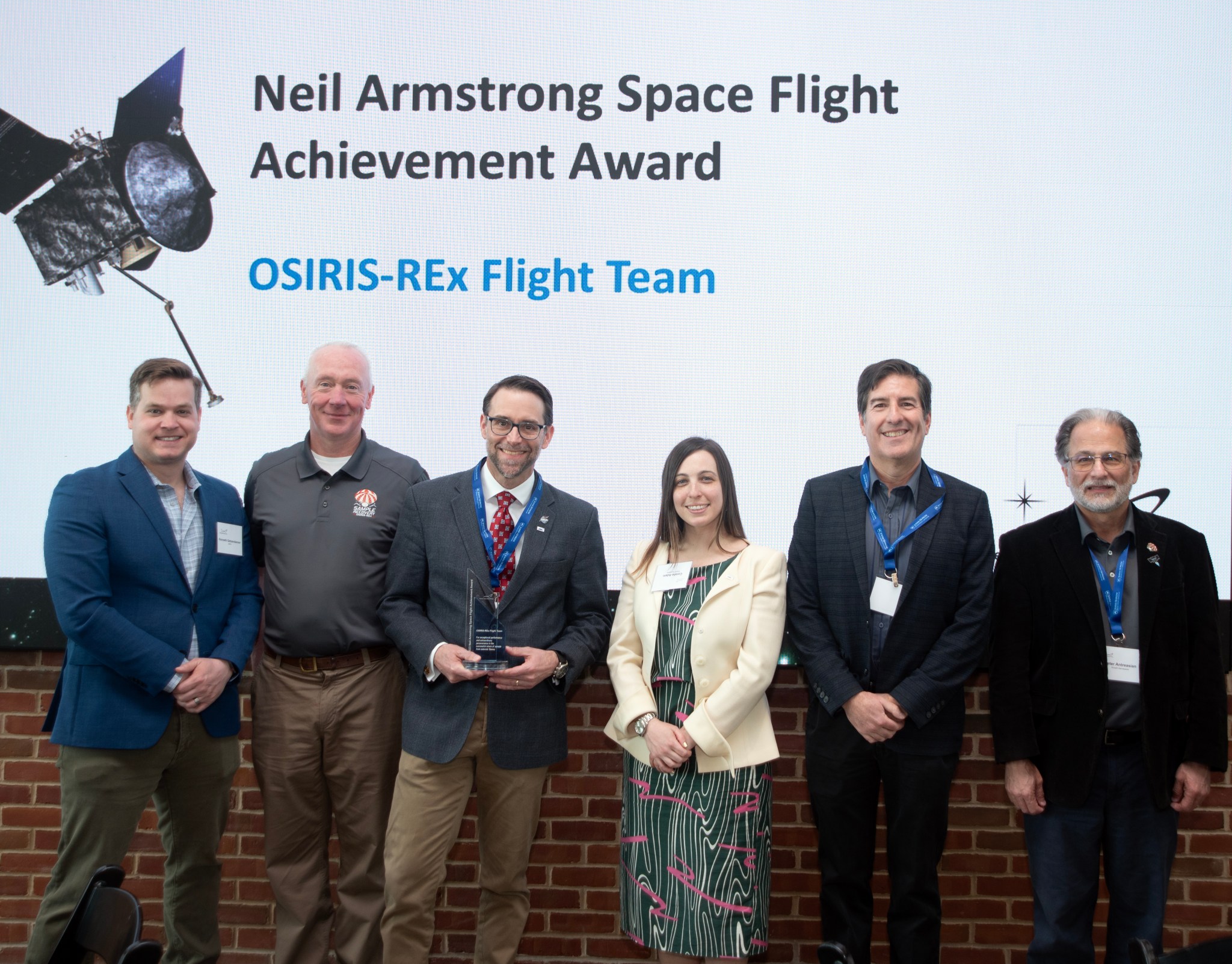 Six members of the OSIRIS-REx team pose for a photo in front of a screen that says: 