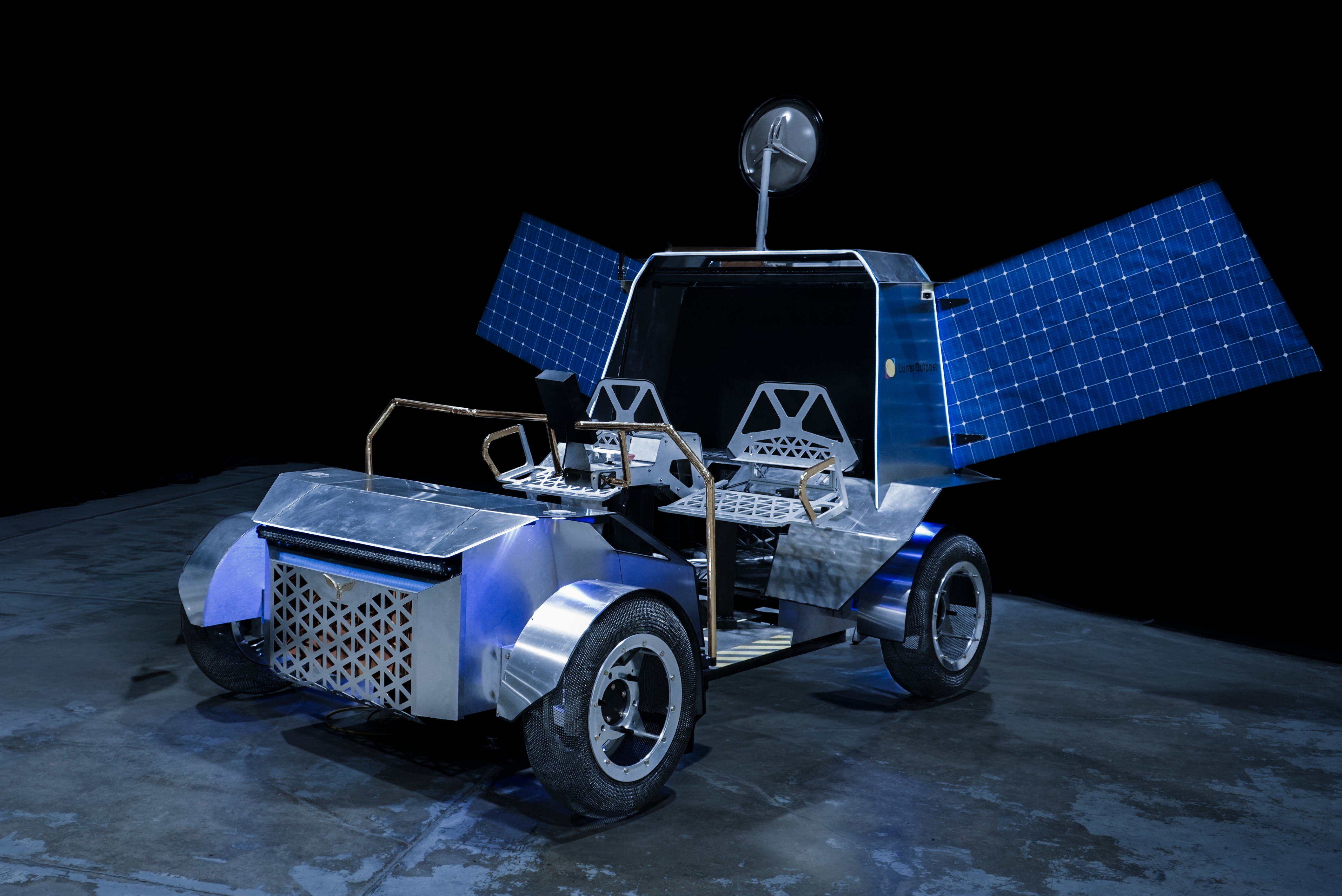 A full-scale prototype lunar terrain vehicle sits on a dark studio floor against a black background. The vehicle has four wheels, two crew seats, deployed blue solar arrays extending from both sides, and a communications dish mounted above the rear section.