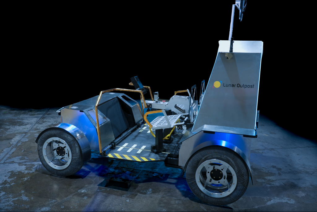 A full-scale prototype lunar terrain vehicle sits on a dark studio floor against a black background. The vehicle has four wheels, two crew seats, deployed blue solar arrays extending from both sides, and a communications dish mounted above the rear section.