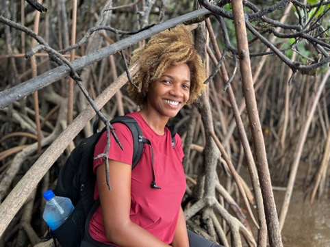 A woman is centered in the image seen from the waist up and is wearing a bright red colored short. She also has a black backpack on her back. Her body is facing towards the right side of the image while her head is facing the camera. Surrounding and behind the woman are several branches, criss-crossing in different directions.