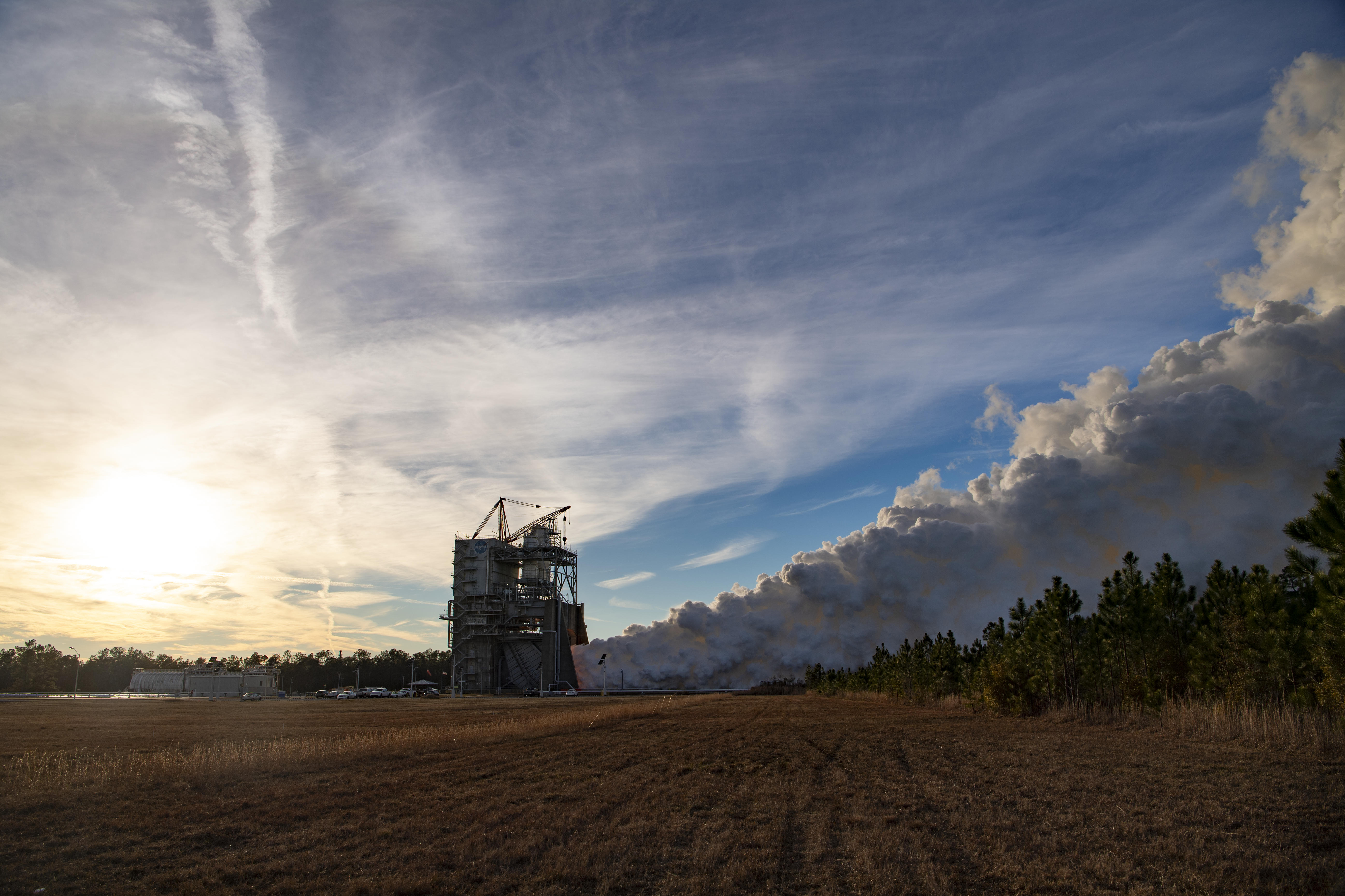 an RS-25 engine test is seen across a grassy field; the sun is visible behind the clouds
