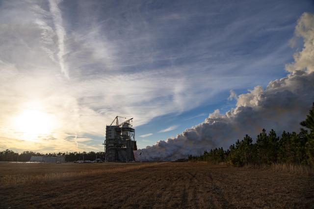 an RS-25 engine test is seen across a grassy field; the sun is visible behind the clouds