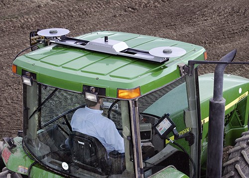 RTK AutoSteer GPS system antennas roof-mounted on a green farming tractor, operated by a man in a blue shirt.