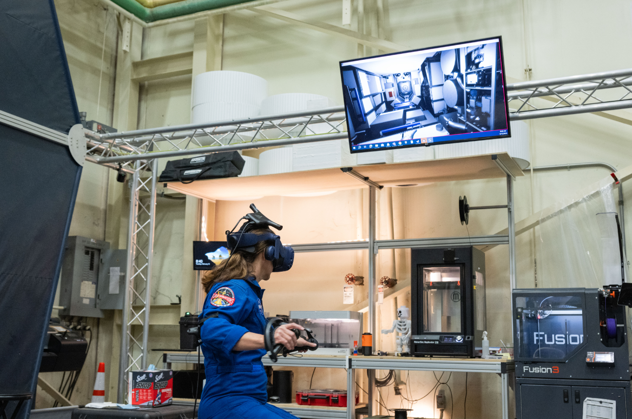NASA Astronaut Nicole Mann wearing a VR headset and holding VR controllers in both hands, immersed in training at the Virtual Reality Training Lab at NASA's Johnson Space Center.