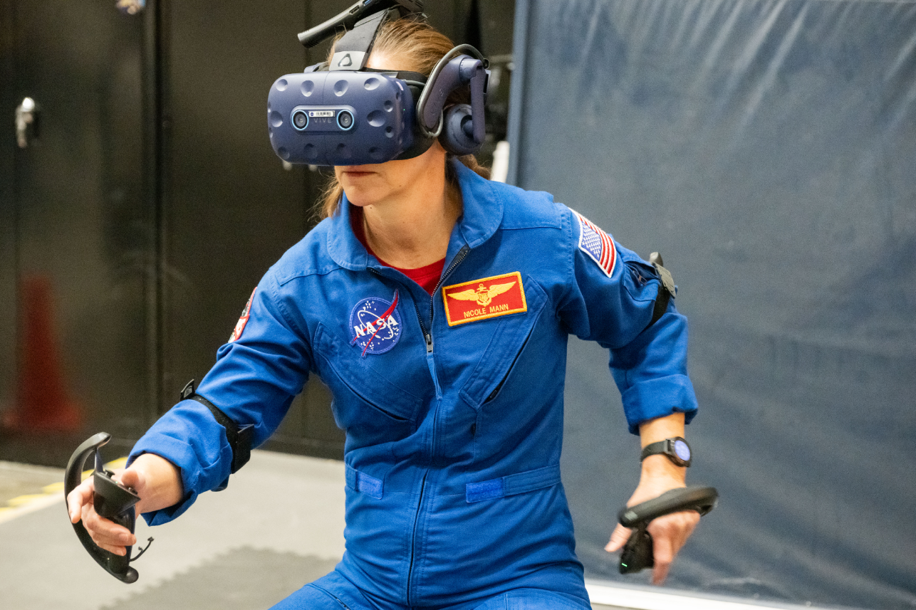 NASA Astronaut Nicole Mann wearing a VR headset and holding VR controllers in both hands, immersed in the simulation at NASA's Johnson Space Center.