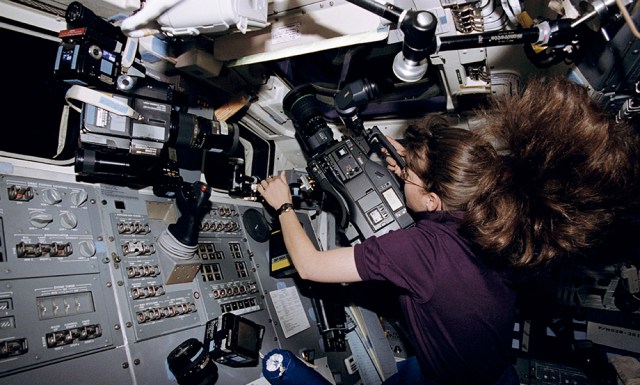 An astronaut aboard a space shuttle points a video camera out the window.