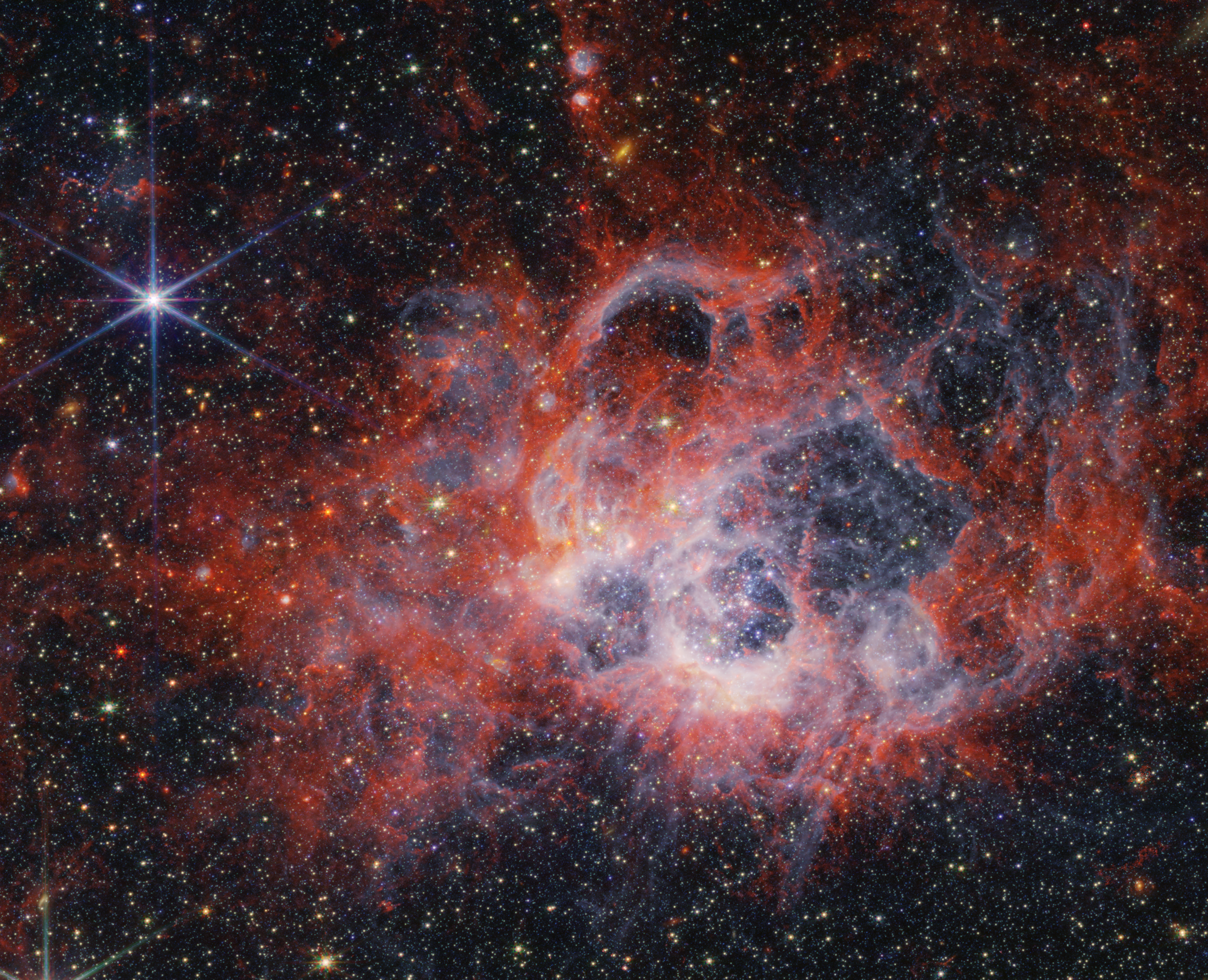 At the center of the image is a nebula on the black background of space. The nebula is comprised of clumpy, red, filamentary clouds. At the center-right of the red clouds is a large cavernous bubble, and at the center of the bubble there is an opaque blueish glow with speckles of stars. At the edges of the bubble, the dust is white. There are several other smaller cavernous bubbles at the top of the nebula, including two tiny cavities at the top center of the image. There are thousands of stars that fill the surrounding area outside the nebula, most of them are yellow or white. At 11 o’clock and 6 o’clock there are extremely bright stars with 8 diffraction spikes. There are also some smaller, red stars and a few disk-shaped galaxies scattered across the image.