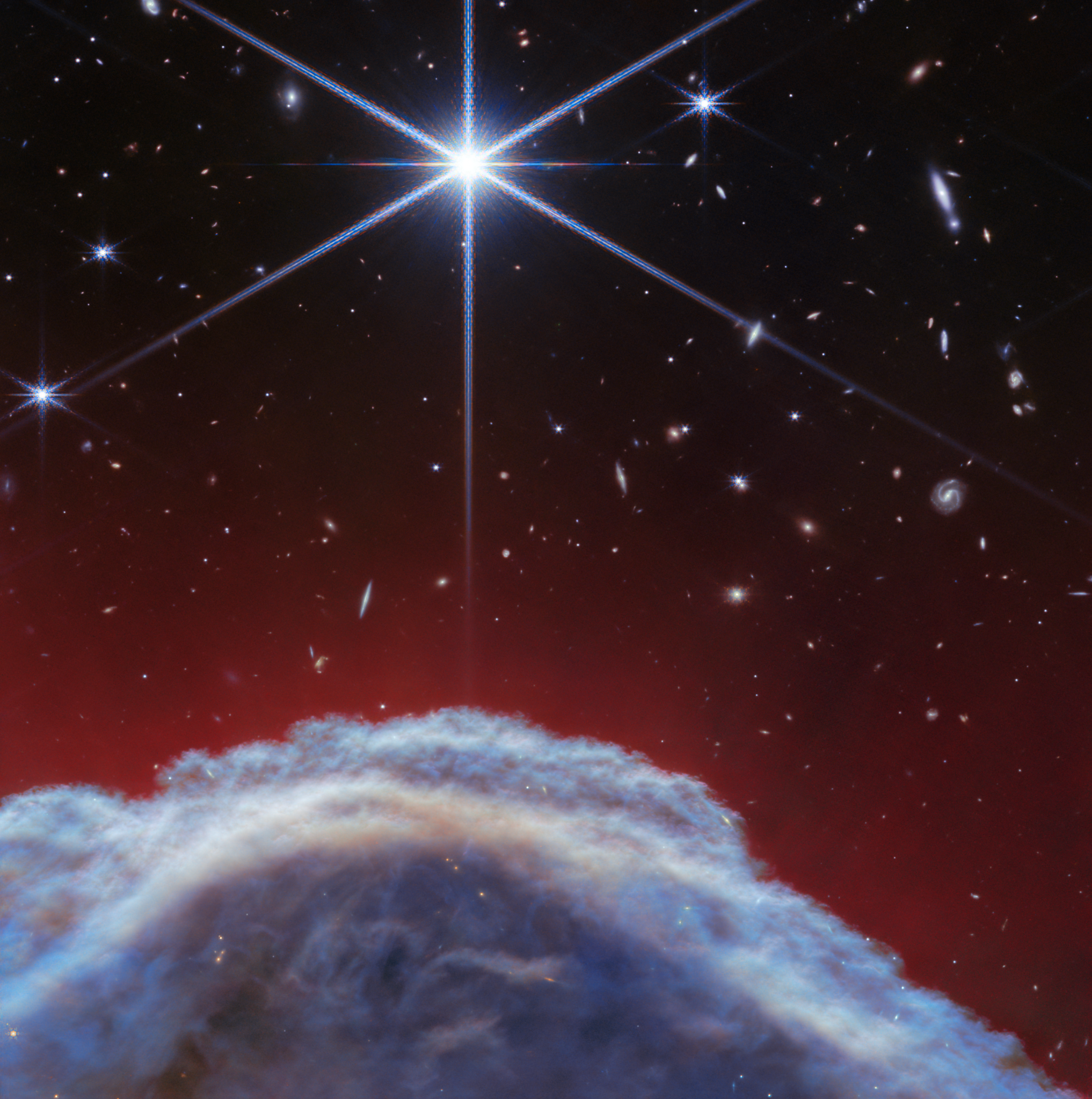 A clumpy dome of blueish-gray clouds rises about a third of the way from the bottom. Above it, streaky, translucent red wisps brush upward to about halfway up the image. The top half of the image is the black background of space with one prominent, bright white star with Webb’s 8-point diffraction spikes. Additional stars and galaxies are scattered throughout the image, although very few are seen through the thick clouds at bottom, and all are significantly smaller than the largest star.