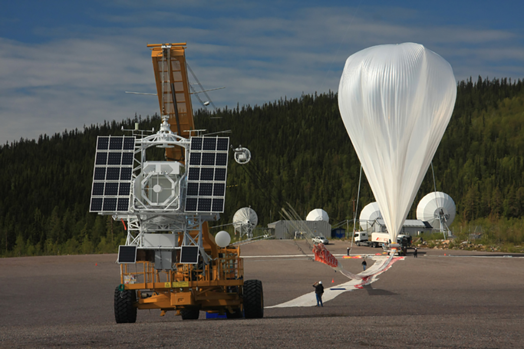 A large metal scientific instrument with black panels is suspended off the ground by a yellow crane. A white trail of material is on the ground and attaches to a partially inflated white balloon in the background.