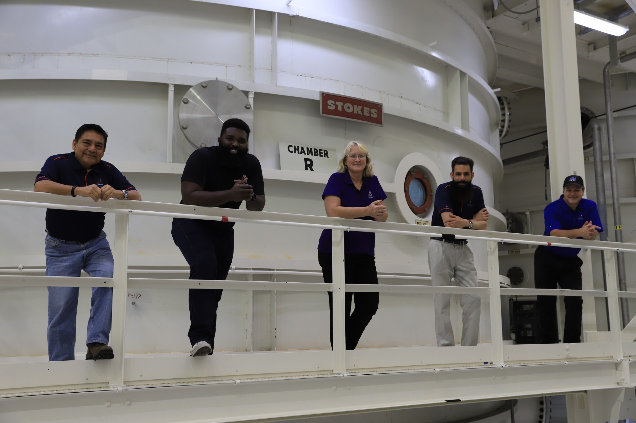 Five people stand in a horizontal line on an elevated platform in front of aa large metal chamber.