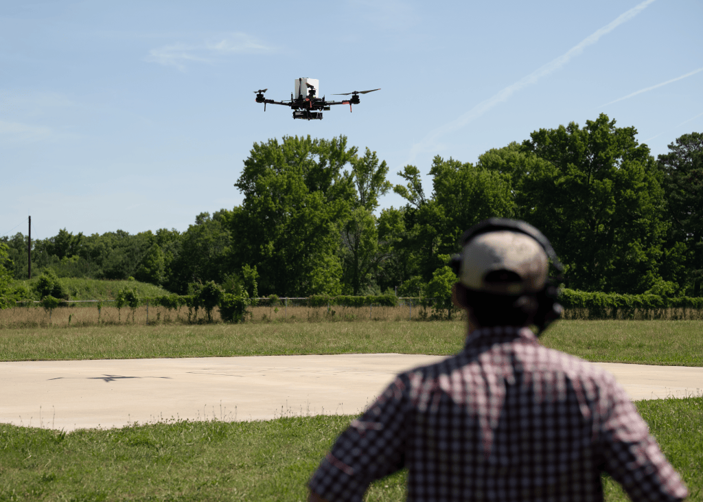 A drone flies over a grassy field at NASA’s Langley Research Center in Virginia as a researcher wearing a headset watches from the foreground. The flight is part of NASA and UNOS research exploring drones for faster, more reliable transport of donor organs.