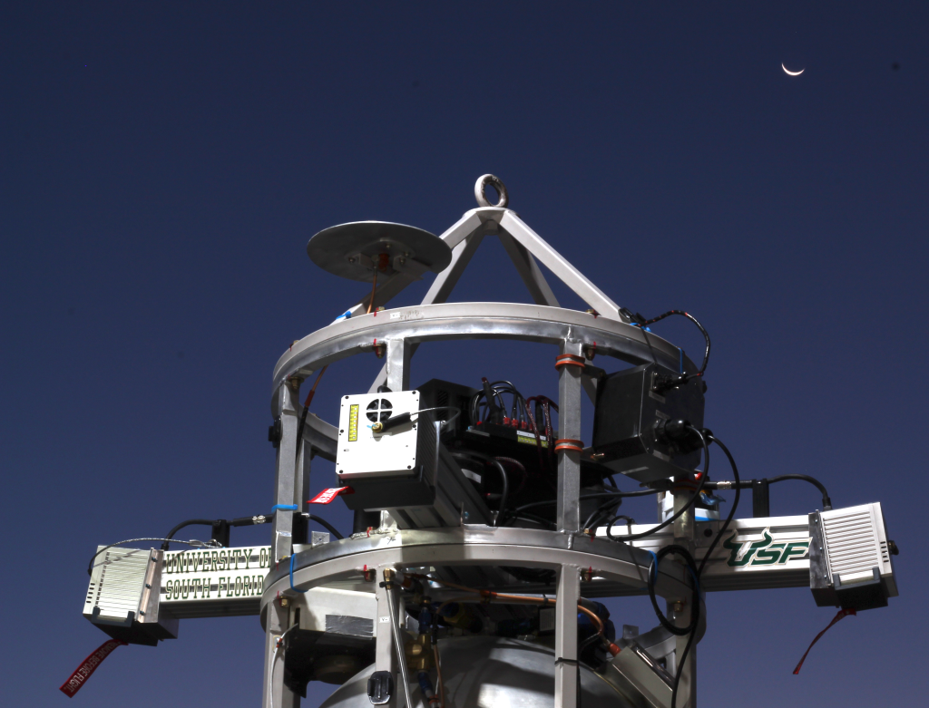 A cylindrical metal frame with a pointed conical top and four rectangular extensions (lidar sensors) jutting from each quadrant houses a payload.