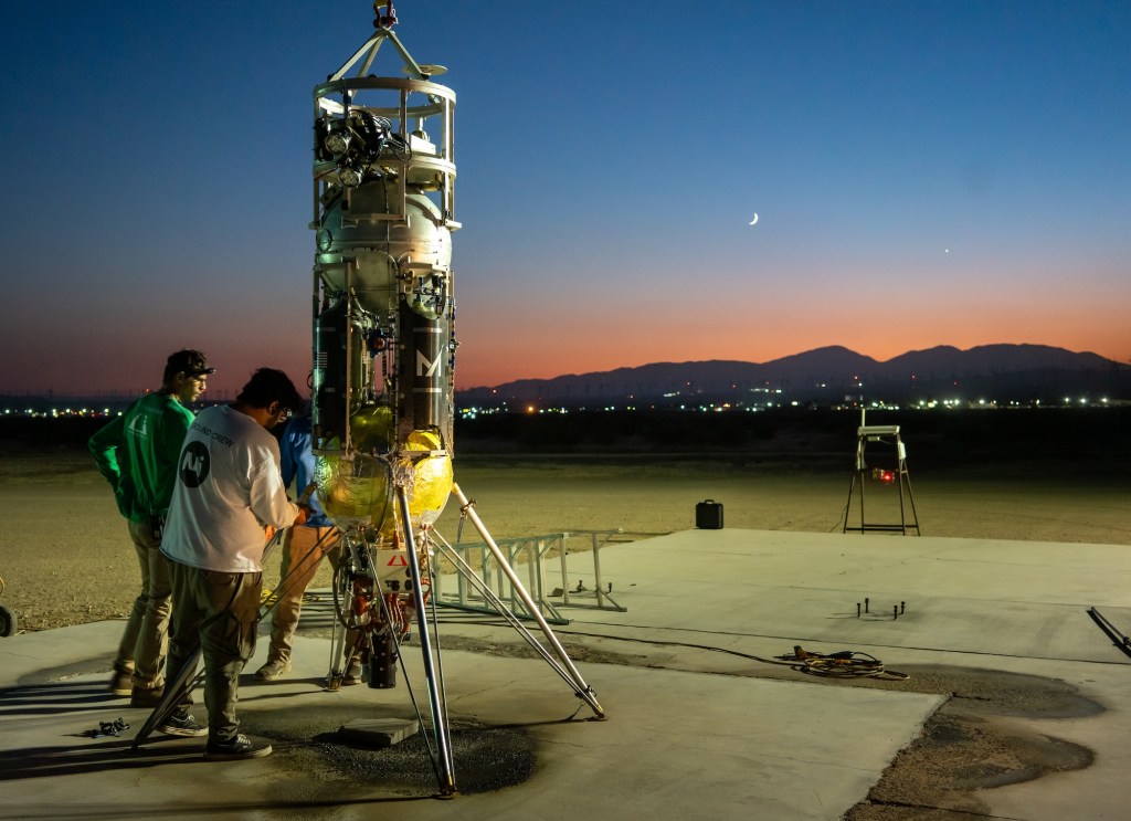 Three researchers make adjustments to a rocket sitting on a launchpad. City lights and the glow of a sunset are in the background.