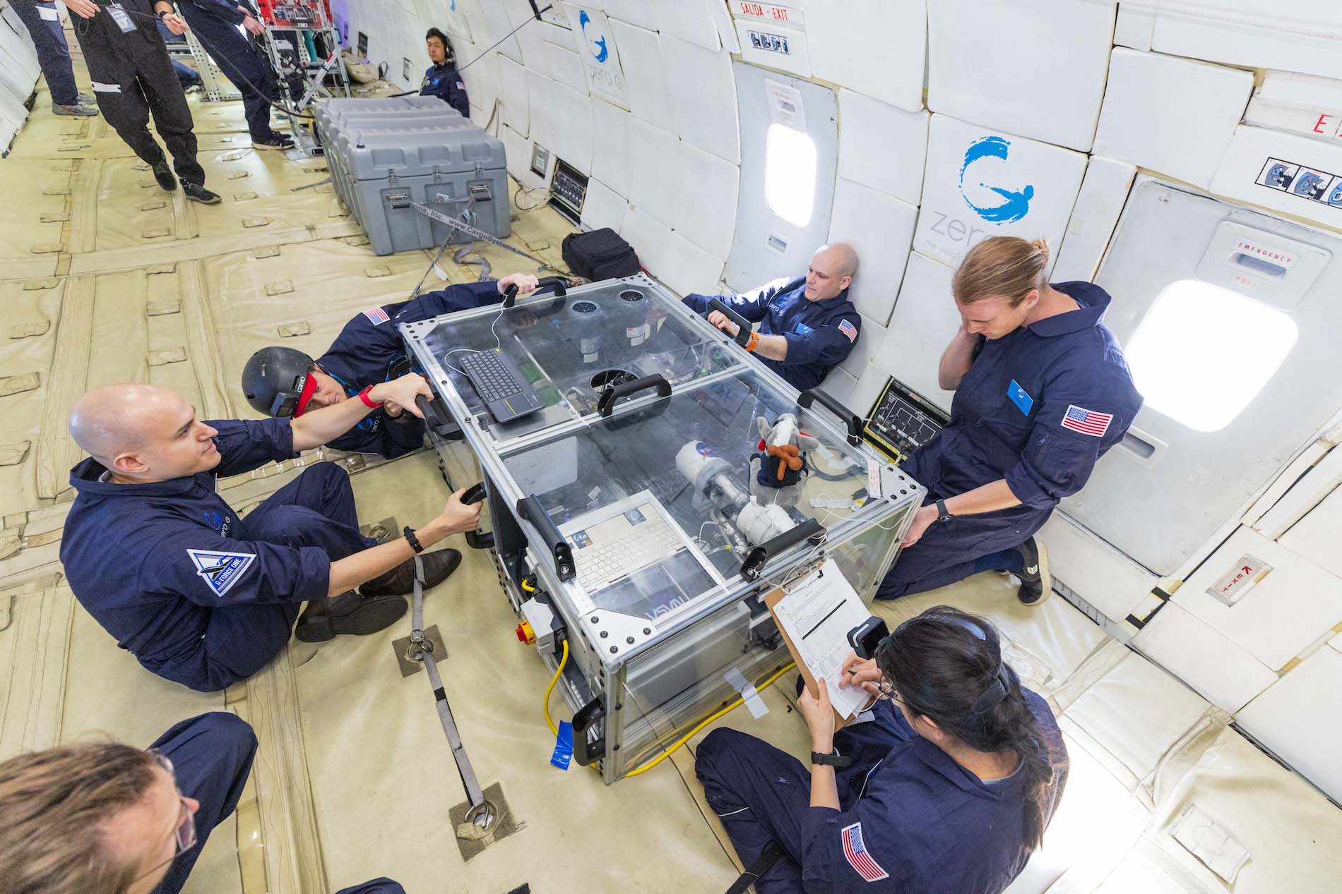 Six researchers in blue jumpsuits on a parabolic flight study their experiment housed in an acrylic and metal frame. One researcher is writing on a clipboard.