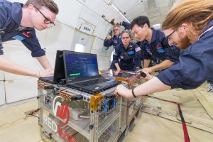 Four researchers float in zero gravity on a parabolic flight while holding onto their experiment encased in a clear case.
