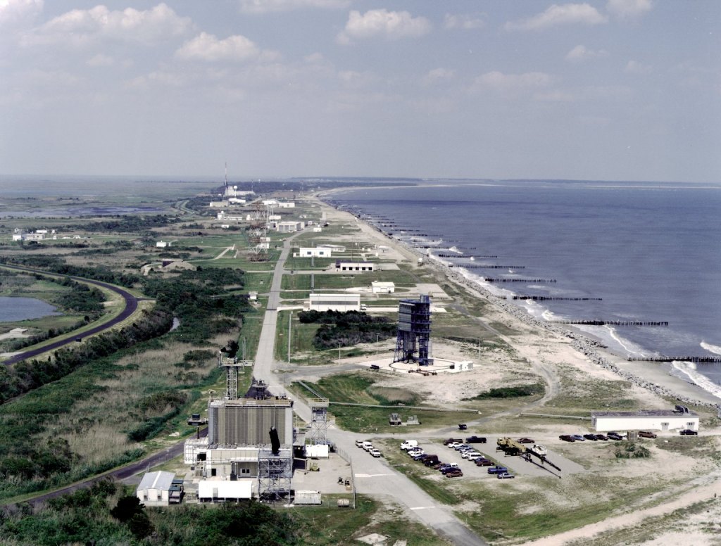 1982 image of a sounding rocket launcher and vertical rocket launcher with support buildings and roads on Wallops Island, Virginia. Ocean groins visible to the east.